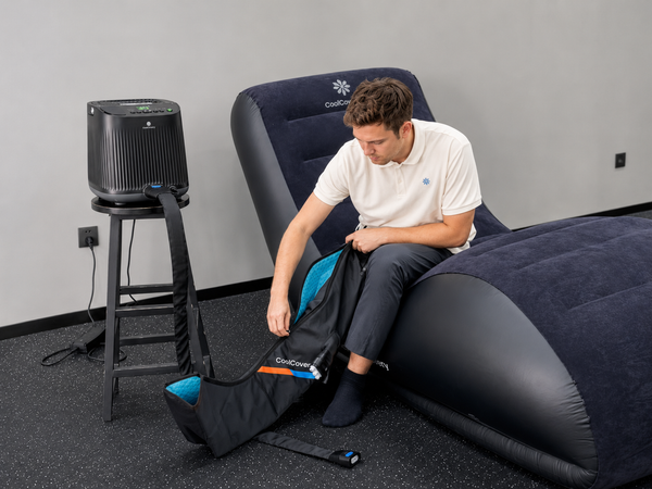 A man sits on a dark navy inflatable lounge chair while unpacking a black branded bag containing an electric massager. A black air pump sits on a stool to the left in a clean indoor room.